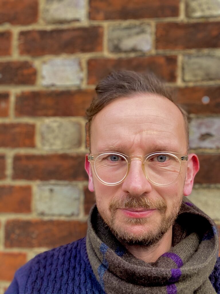 Dan Hicks, Professor of Contemporary Archaeology at the University of Oxford [photograph against a brick wall, Professor Hicks is wearing a blue knitted jumper, with a grey scarf and clear-framed glasses]